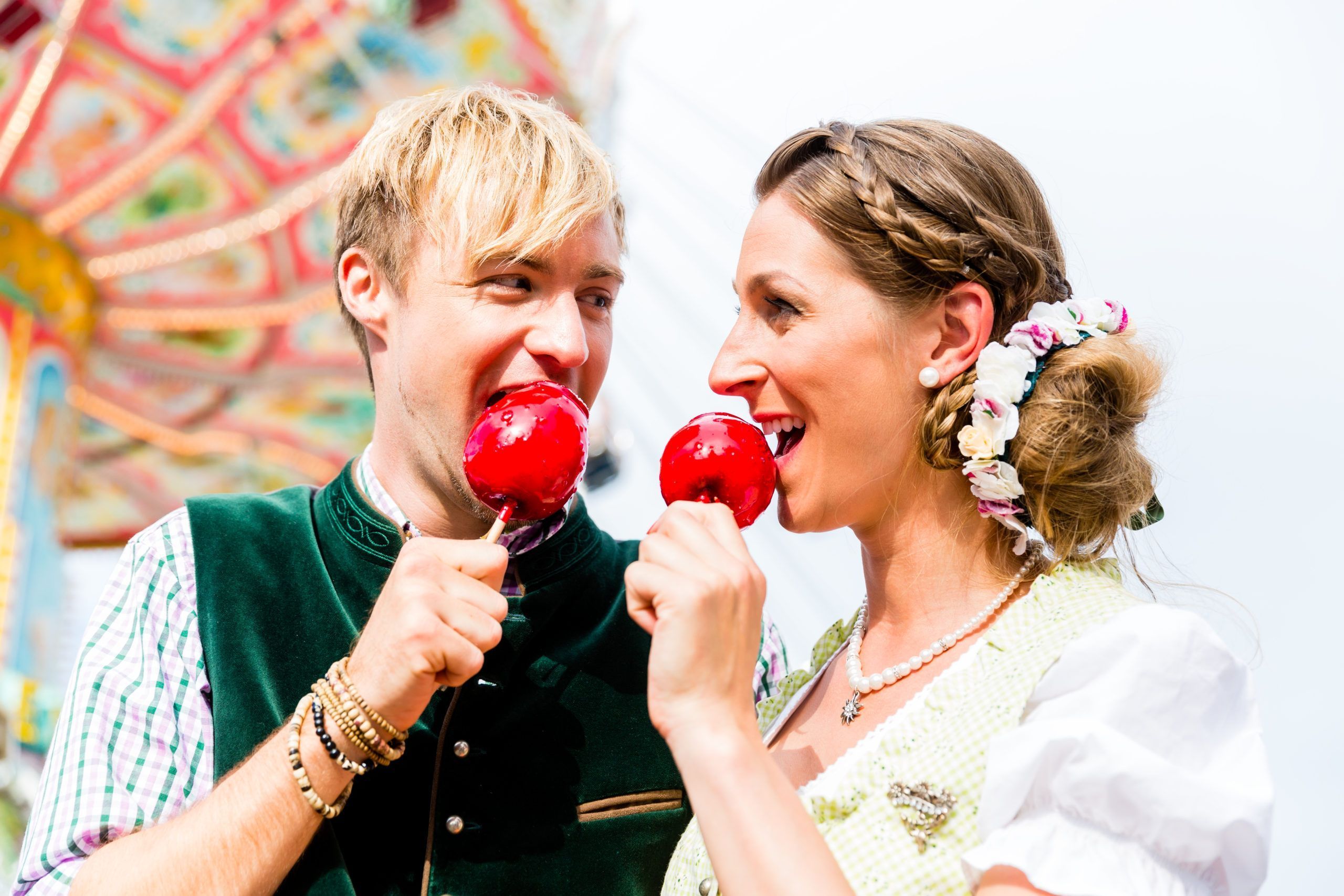 Woman and man biting red candy apples wearing Bavarian traditional clothes in front of carousel