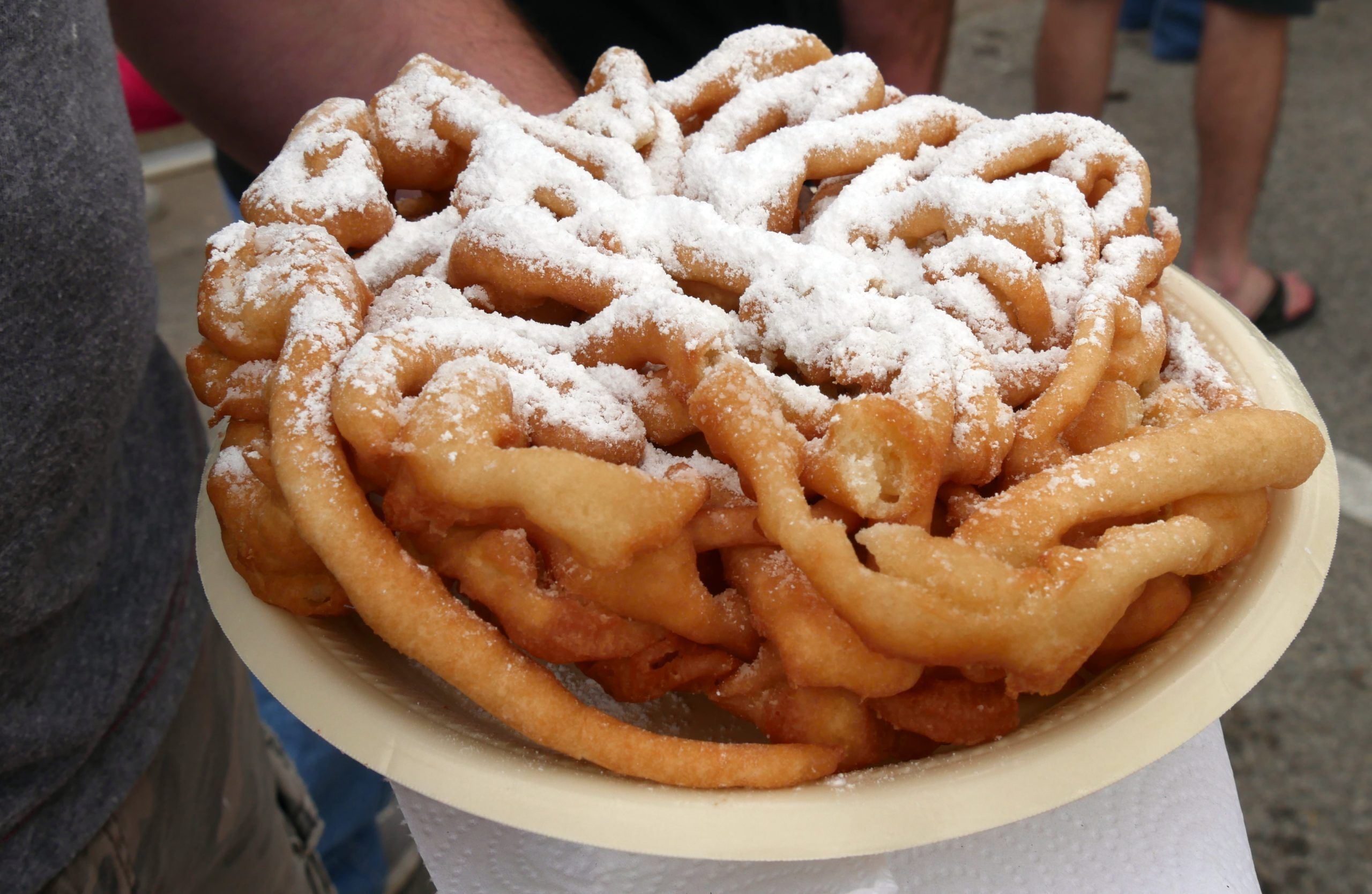 A hand holding a plate of funnel cake with powdered sugar on top