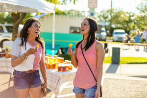 Two women at the Colorado State Fair wearing summer garb laugh while enjoying a snack and walking the midway