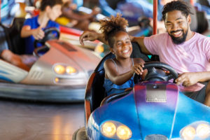 A little girl smiles and steers a bumper car with her dad sitting next to her at the Colorado State Fair