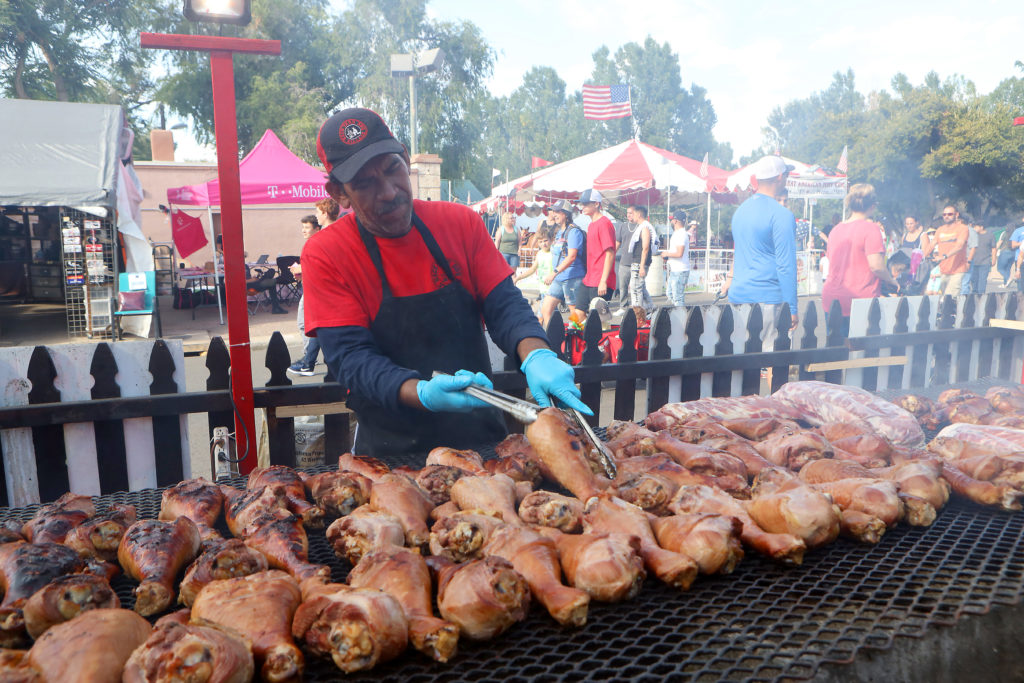A man in an apron tends an outdoor grill filled with turkey legs at the Colorado State Fair
