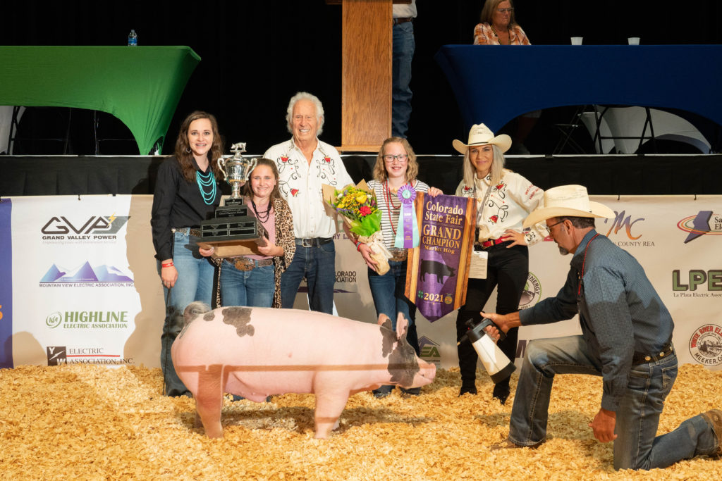 Two teen girls stand holding banners and trophies along with several other people showing that their pic standing in the foreground won Grand Champion Market Hog