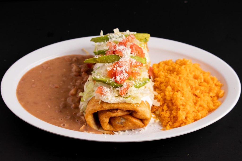 A chimichanga garnished with cheese and peppers sits on the middle of a plate with rice on one side and refried beans on the other